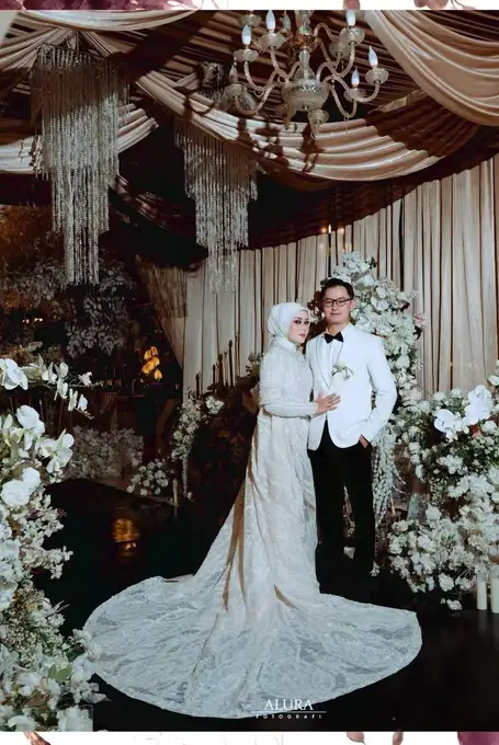 Pengantin berbusana putih berdiri di antara rangkaian bunga putih dan lampu kristal.