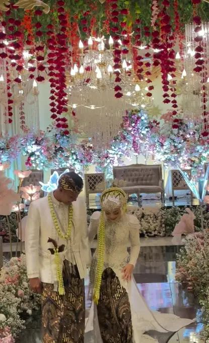 Pengantin berjalan bersama di depan pelaminan dengan untaian bunga merah.