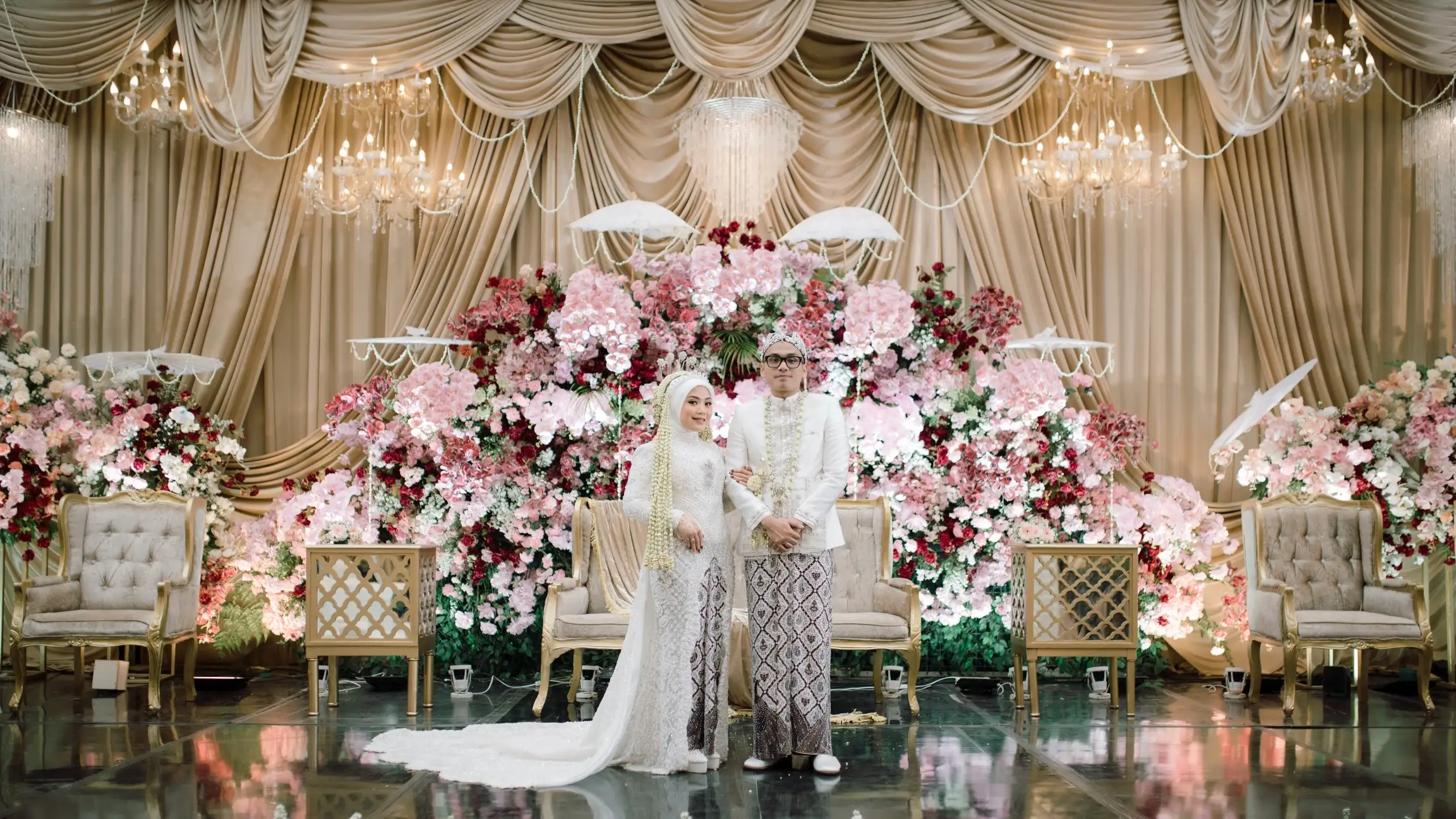 Pasangan pengantin berdiri di pelaminan yang dipenuhi bunga merah muda.