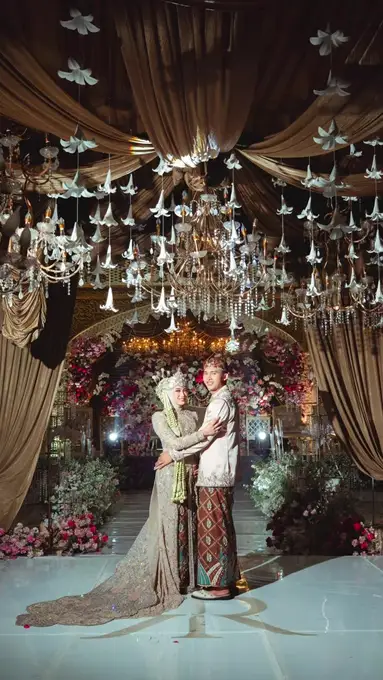 Pengantin berbusana adat berpelukan di bawah langit-langit lampu gantung dan tirai cokelat.