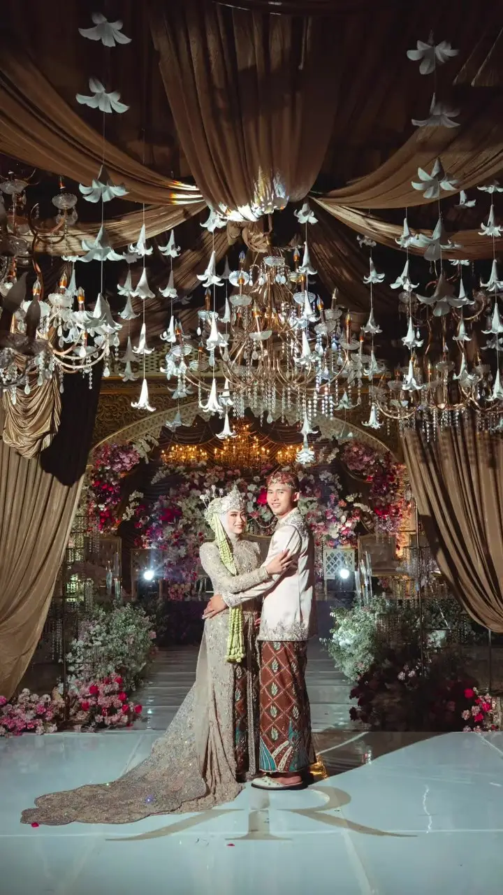 Pengantin berbusana adat berpelukan di bawah langit-langit lampu gantung dan tirai cokelat.
